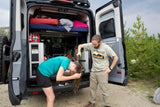 Man helps woman wash hair with an outdoor shower from their RV rental van.