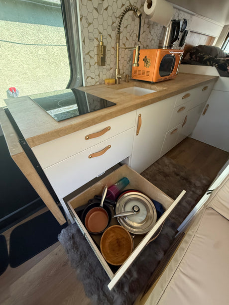 Cozy, compact rv rental kitchen with a modern cooktop, sink, retro microwave, and open drawer showing cooking essentials.