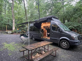 Grey camper van with open door, showing wooden interior, parked under an extended awning at a forest campsite. Perfect rv rental.