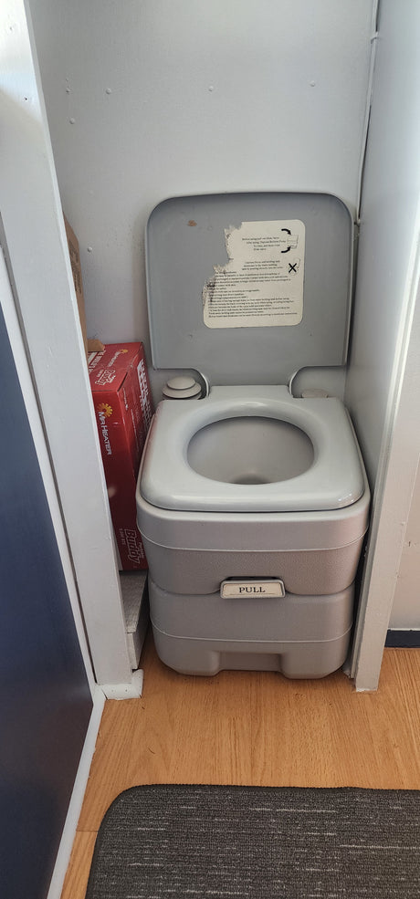 A grey portable toilet, commonly found in an RV rental, sits in a small washroom alongside a red heater box.