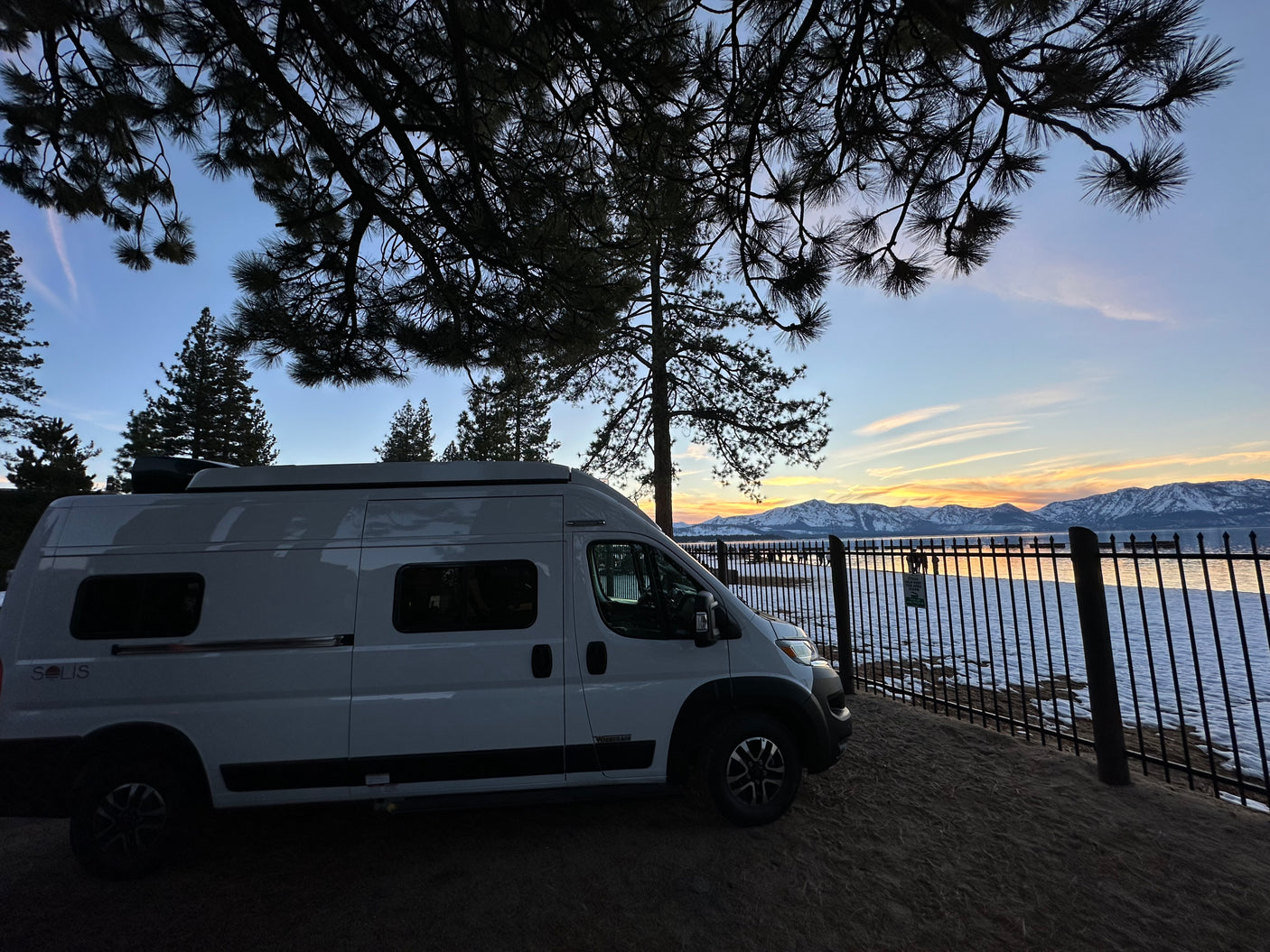 An RV rental camper van parked by a lake with snowy mountains reflecting sunset hues, framed by silhouette pine trees.