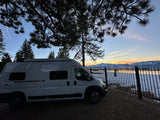 An RV rental camper van parked by a lake with snowy mountains reflecting sunset hues, framed by silhouette pine trees.