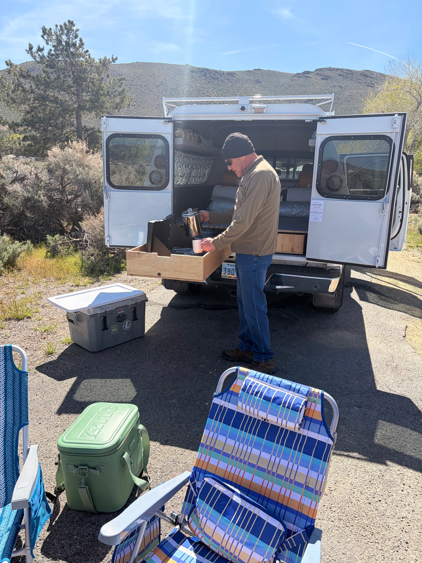 Man pours coffee from an RV rental camper van, enjoying morning at a scenic campsite. Coolers & chairs ready for adventure.