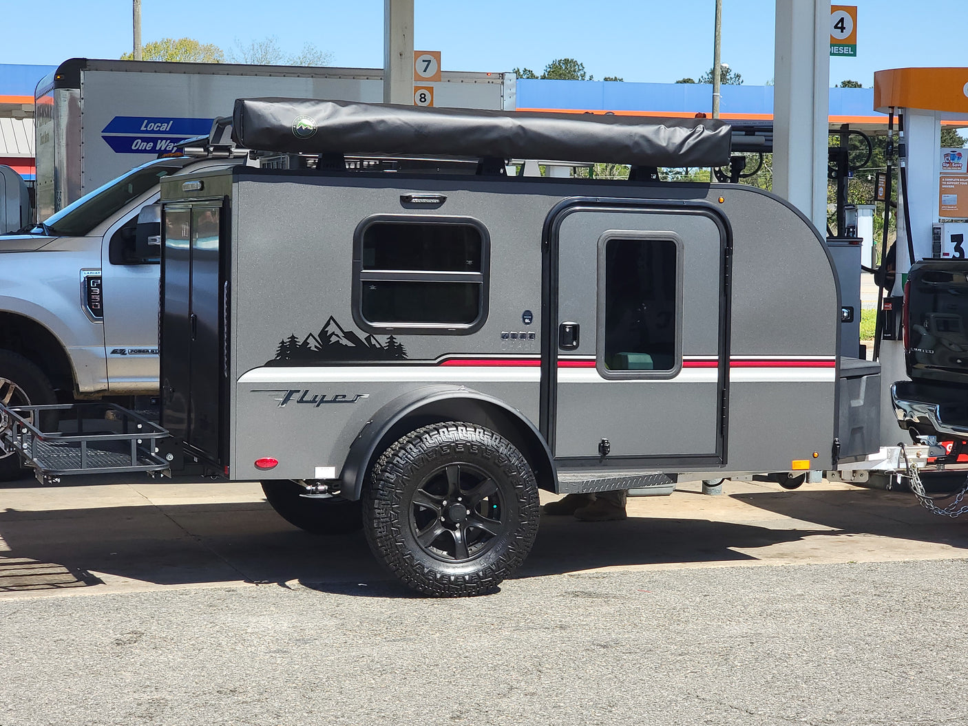This grey compact RV rental trailer, pulled by a Ford truck, is fueling up at a gas station for its next adventure.