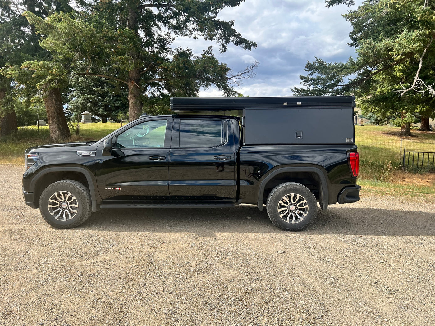 Black GMC Sierra AT4 truck with a black truck bed camper for an RV rental. Parked on gravel beside trees & a cemetery. — photo 5