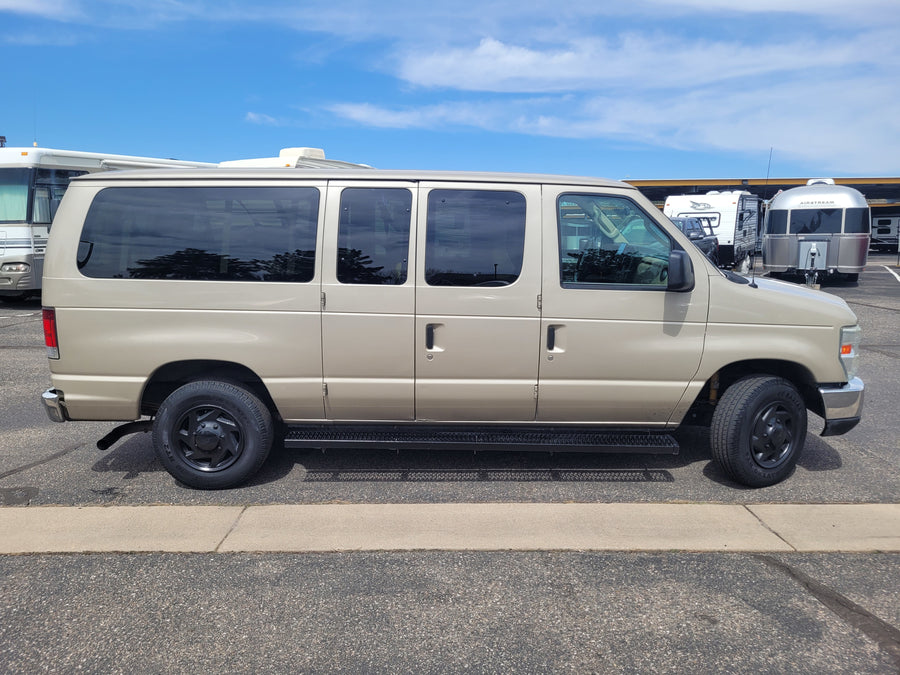 This tan Ford van is parked among various RVs, including an Airstream trailer, possibly for an RV rental business.