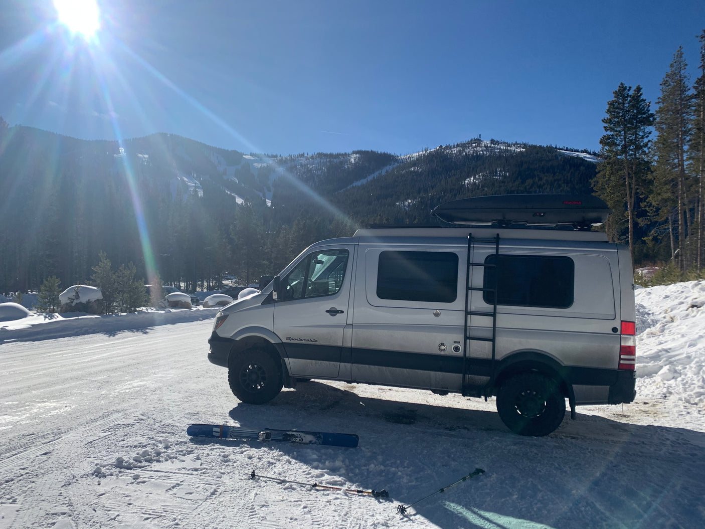 A silver RV rental van is parked in snowy mountains with skis on the ground, bathed in bright sunlight.