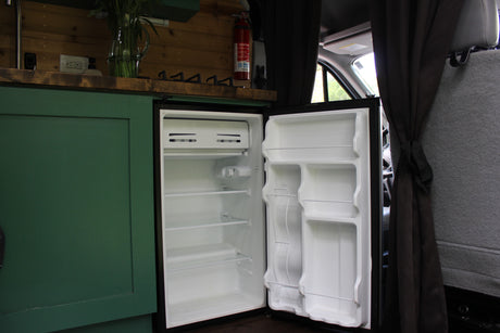 Empty mini-fridge in an RV rental camper van, showing shelves & door storage. Adjacent to a green cabinet, part of the vehicle interior.