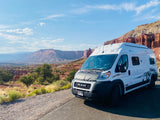 A white camper van RV rental overlooks stunning red rock desert canyons and mesas under a clear blue sky.