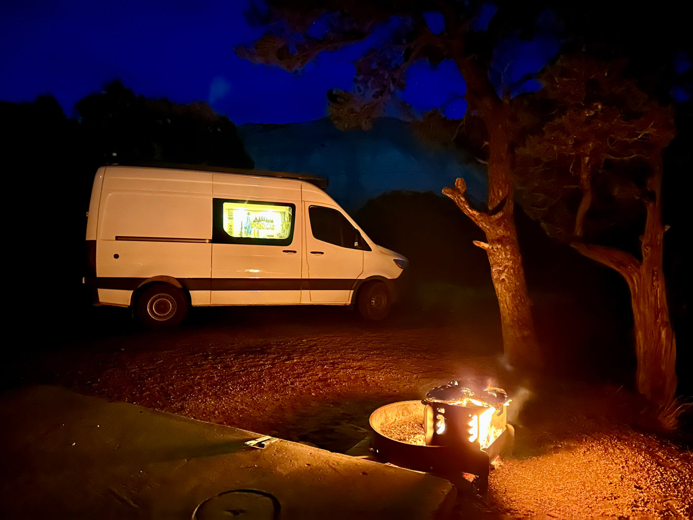 Night scene with a white RV rental, fire pit, and trees under a dark blue sky, creating a cozy camping atmosphere.