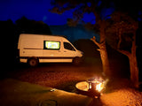 Night scene with a white RV rental, fire pit, and trees under a dark blue sky, creating a cozy camping atmosphere.