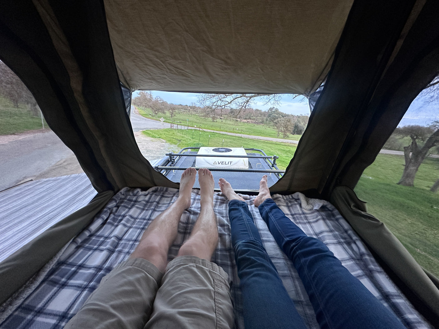 Two people relax in an RV rental rooftop tent with feet up, enjoying a scenic view with an AVELIT unit on the roof.