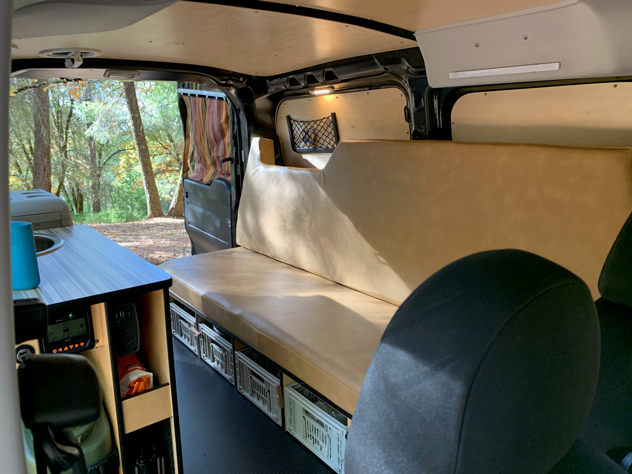 Inside an RV rental camper van: a cozy bench bed, kitchen, storage crates, and forest views from the open door.