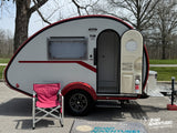 This T@B 320 teardrop RV rental, white with red trim, is parked outside with a camping chair, ready for adventure.