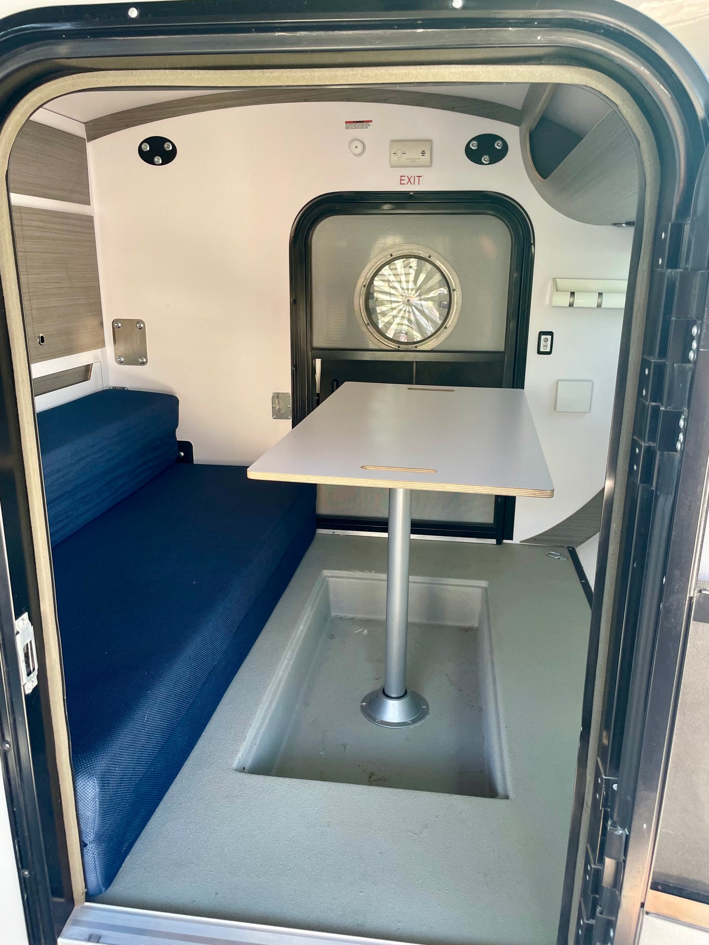 Inside a compact RV rental: blue bench seating, a central table, and an exit door with a round window.