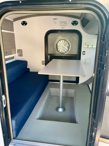 Inside a compact RV rental: blue bench seating, a central table, and an exit door with a round window.
