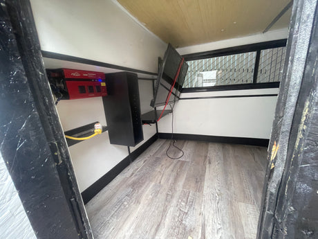 Compact RV rental interior with power inverter, monitor, storage shelves, and a window with a grate.