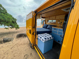 This vibrant yellow RV rental van is parked on a sandy beach, door open to reveal a cozy, wood-paneled interior overlooking the ocean.