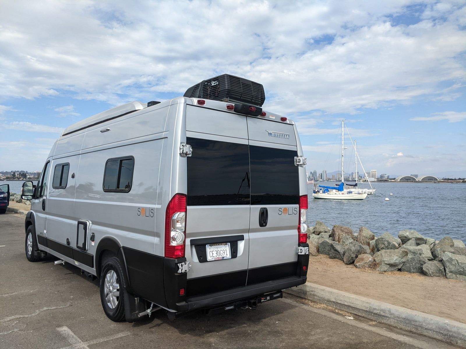 Experience travel with this RV rental! A silver Winnebago Solis camper van parked by the water, with boats and city views.