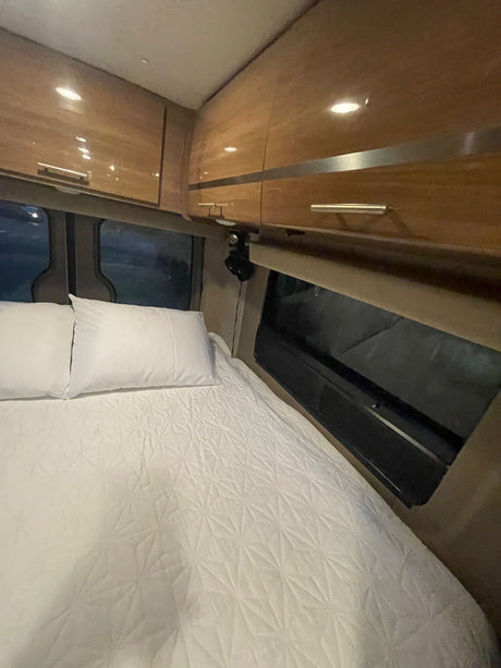 View of a neat RV rental sleeping area: crisp white bedding, pillows, and ample overhead storage.