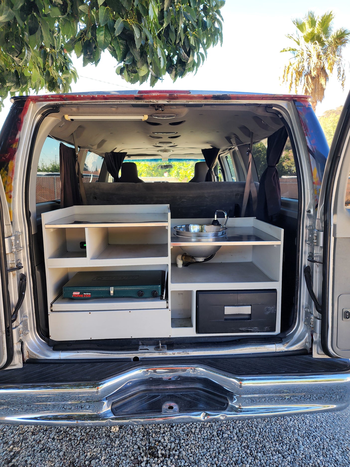 Rear view of a compact campervan interior with a built-in kitchen, sink, stove, and cooler. Perfect for an RV rental adventure!