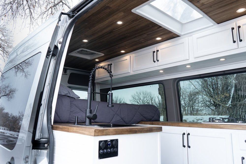 Cozy RV rental interior: Custom camper van features a modern kitchen, comfortable bed, and wood-paneled ceiling.