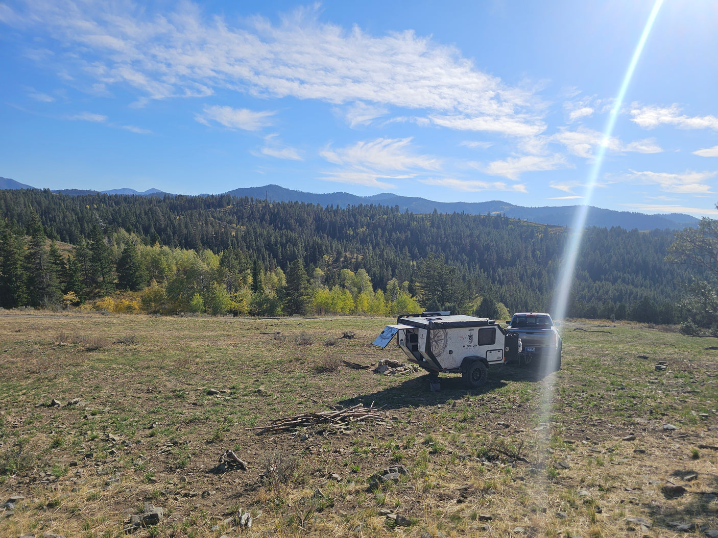 Adventure-ready RV rental trailer and truck parked on a hill overlooking a lush forest and mountains under a sunny sky.