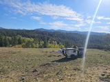 Adventure-ready RV rental trailer and truck parked on a hill overlooking a lush forest and mountains under a sunny sky.