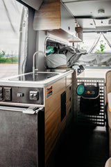 RV rental: Inside a modern camper van. Features a compact kitchen with sink, fridge, controls, and an elevated bed.