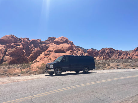 A black van on a desert road by red rock mountains under a bright blue sky. Perfect for an RV rental adventure!