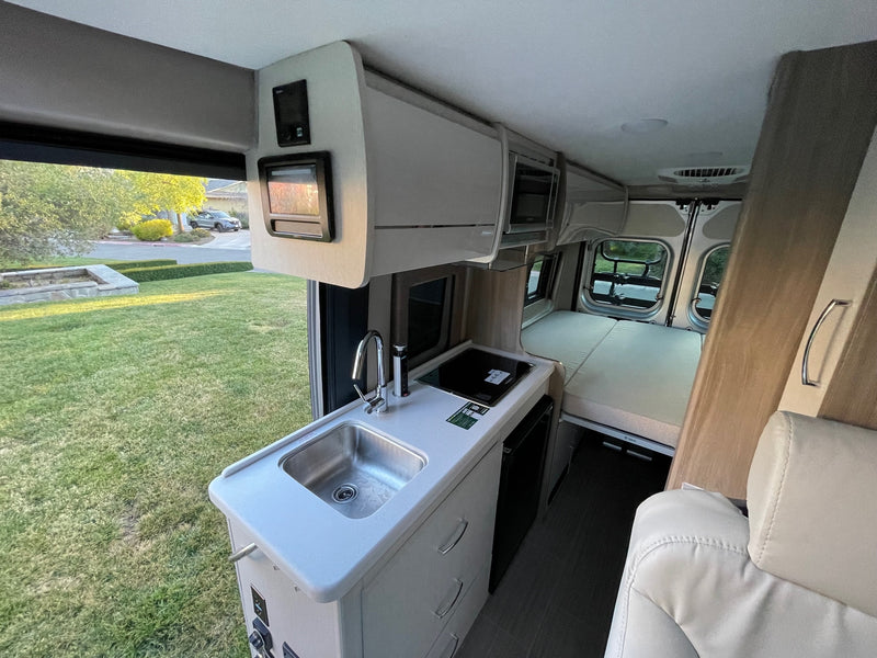 Inside an RV rental, showing the compact kitchen with sink, induction cooktop, rear bed, and outdoor view. — photo 2