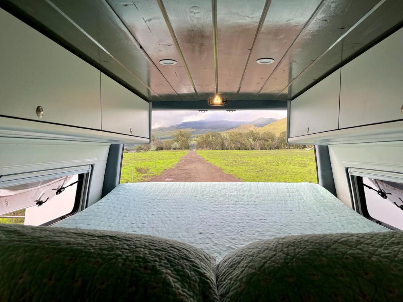 Cozy RV rental bed overlooks a scenic view of a dirt road leading to green fields and mountains.