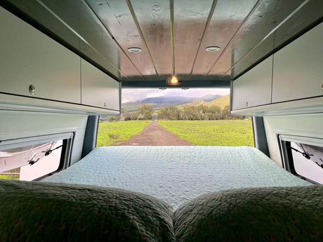 Cozy RV rental bed overlooks a scenic view of a dirt road leading to green fields and mountains.