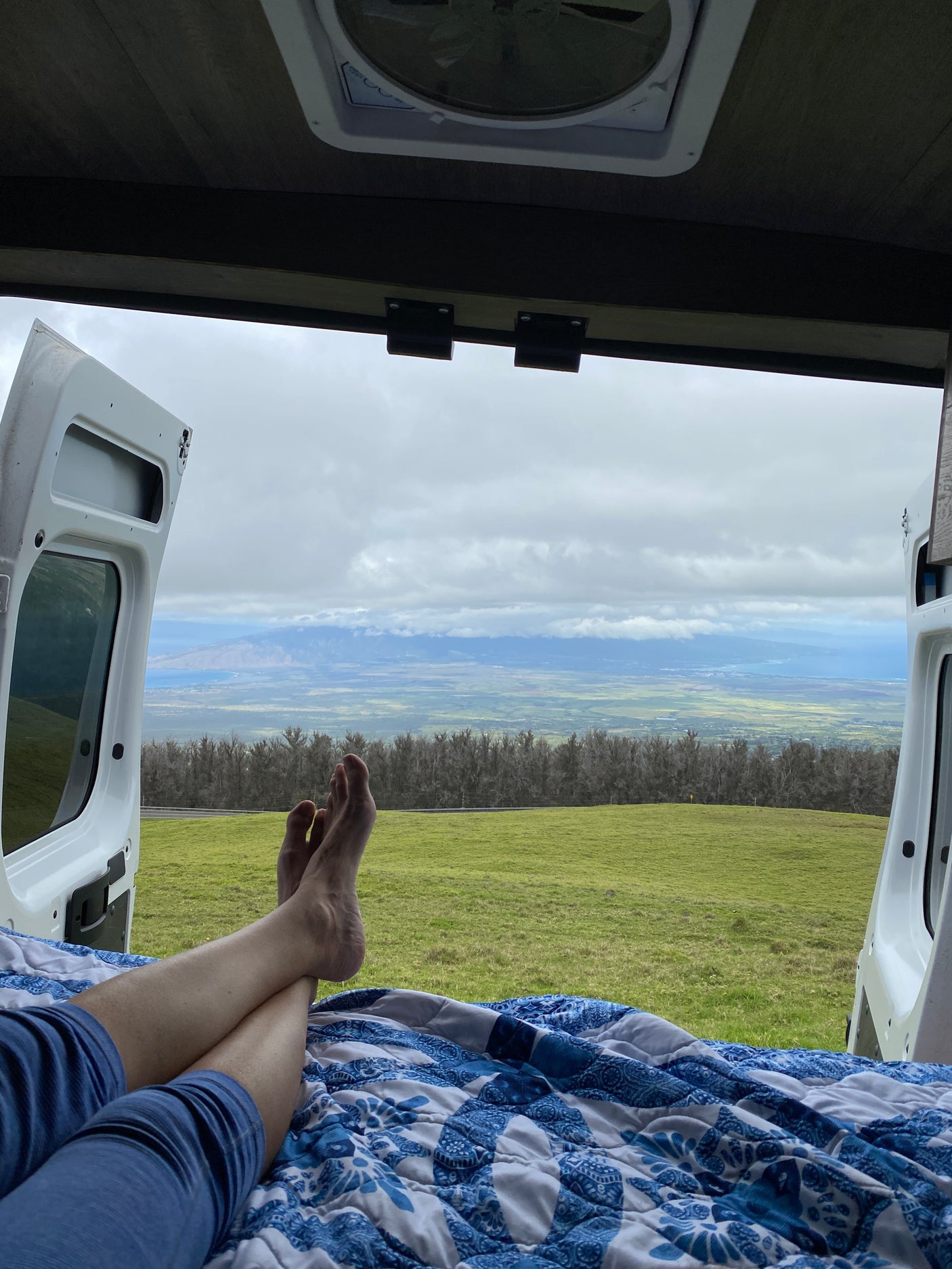 From an RV rental, a person's feet rest on a blue quilt, gazing at a vast green field, distant trees, valley, ocean, and mountains under a cloudy sky.