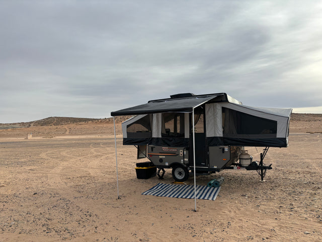 An RV rental pop-up camper is set up for desert camping, with an awning and mat, under an overcast sky.