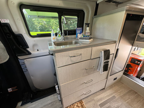 Inside an RV rental, a compact kitchen features a sink, countertop, multiple storage drawers, and a window view of green trees.