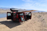 Ready for adventure! A red van with an awning & roof box by a lake & mountains. An ideal RV rental for off-grid camping.