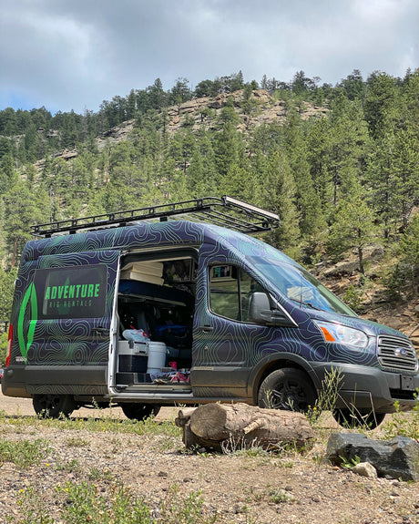 Dark-colored adventure RV rental van with open side door, parked in a forest, showcasing its camper features.