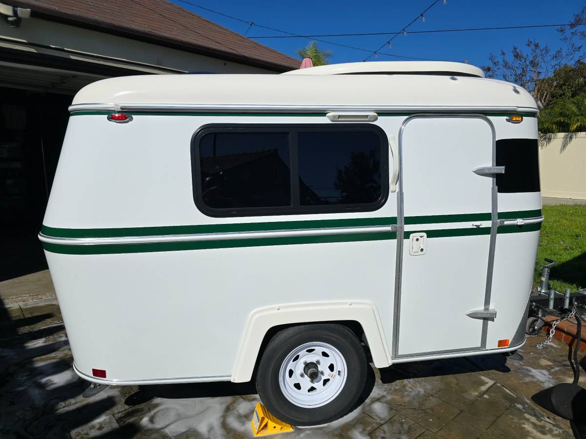  tree-lined campsite for an outdoor adventure.|Side view of a pristine white compact travel trailer with green stripes