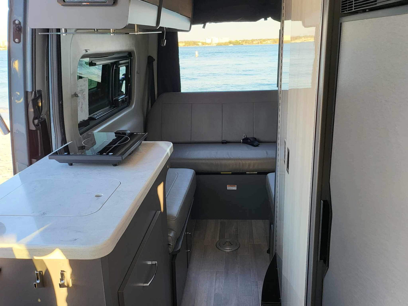RV rental interior: kitchen counter with portable cooktop, bench seat, and a large window overlooking the water.