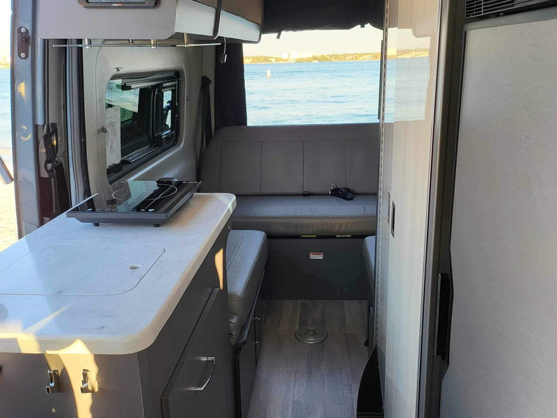 RV rental interior: kitchen counter with portable cooktop, bench seat, and a large window overlooking the water. — photo 12