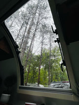 Cozy RV rental view: rain streaks on the window, tall trees, and a parked vehicle in the tranquil forest.