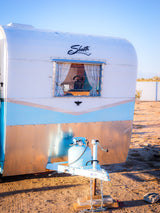 Vintage Shasta RV rental, white and light blue with a wood-look base, parked outdoors with a propane tank visible.