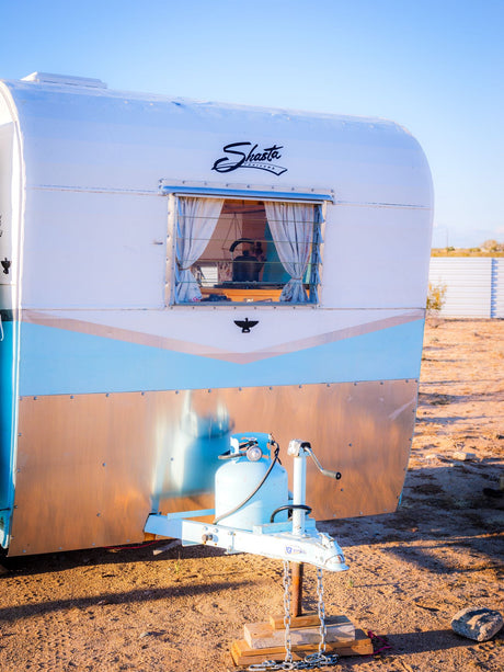 Vintage Shasta RV rental, white and light blue with a wood-look base, parked outdoors with a propane tank visible.