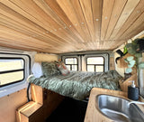 Cozy RV rental interior featuring a wooden plank ceiling with lights, a comfortable bed with pillows, and a compact sink area.