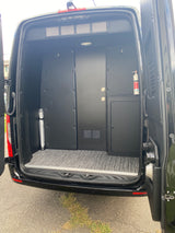View inside a black RV rental van. Custom interior panels, a storage door, safety gear, and patterned flooring.