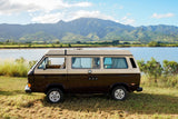Iconic brown/beige RV rental parked by a serene lake, framed by majestic green mountains under a bright sky.