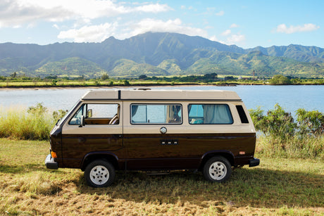 Iconic brown/beige RV rental parked by a serene lake, framed by majestic green mountains under a bright sky.