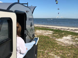 Woman relaxing in a FREESOLO RV rental by the beach, watching kitesurfers and a bridge in the distance.