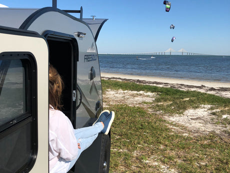 Woman relaxing in a FREESOLO RV rental by the beach, watching kitesurfers and a bridge in the distance.